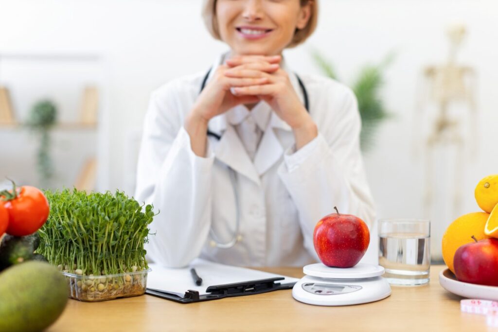 A dietitian smiles while sitting at a table with fresh fruits and vegetables, promoting healthy eating habits