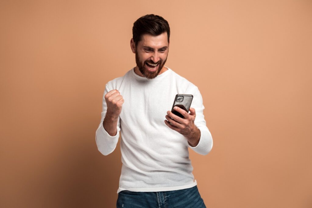 Happy overjoyed man with beard looking at the smartphone and smiling making yes gesture, celebrating victory